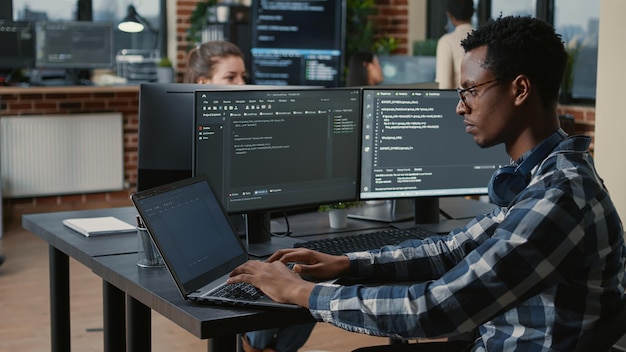 Retrato de un programador cansado frotándose los ojos mientras escribe en una laptop sentada en el escritorio con múltiples pantallas analizando código en una agencia de software. El codificador pierde el foco mientras trabaja en la interfaz de usuario.