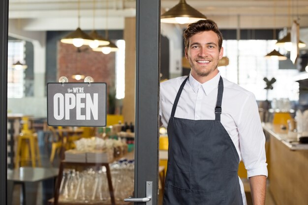 Retrato del propietario sonriente de pie en la puerta de su restaurante con letrero abierto