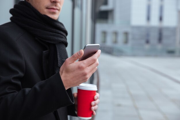 Retrato recortado de jovem empresário em roupas quentes com café e telefone ao ar livre