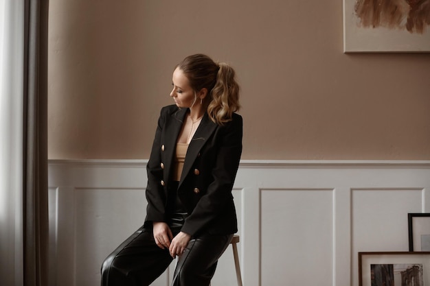 Retrato recortado de uma jovem elegante com maquiagem da moda usando um blazer estiloso e calça preta posando na sala de estar