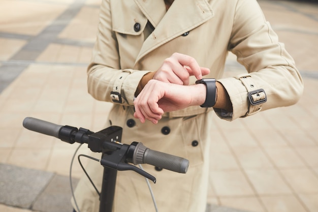 Retrato recortado del hombre de negocios irreconocible con gabardina y control de tiempo mientras viaja en scooter eléctrico para ir al trabajo en el entorno urbano de la ciudad, espacio de copia