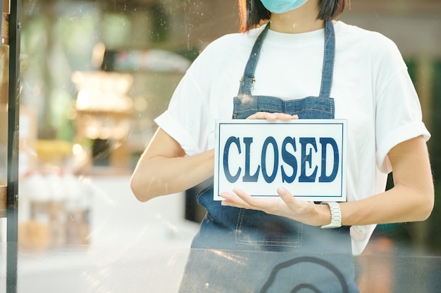 Retrato recortado de mujer barista sosteniendo un cartel cerrado cuando está parado en la entrada de la cafetería