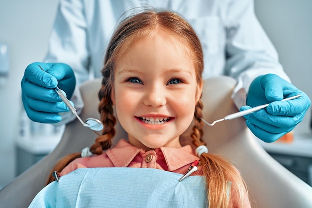 Retrato recortado de una niña con pelo de coletas sentada en un sillón dental mirando la cámara y sonriendo Detrás de un médico con guantes sostiene herramientas de examen Odontología infantil