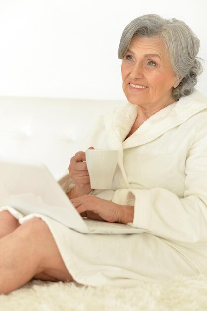 Retrato de relajado seniour mujer trabajando en la computadora portátil y bebiendo té en casa