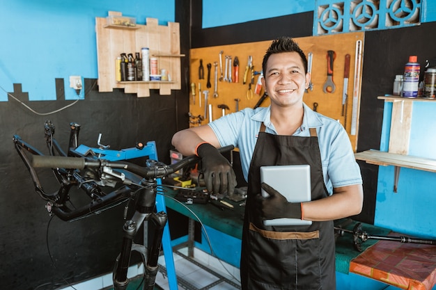 Retrato de un reparador en un delantal apoyado en un cuadro de bicicleta mientras sostiene una tableta