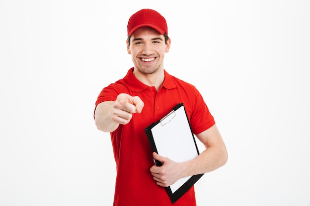 Retrato de repartidor feliz en camiseta roja y gorra sonriendo y señalando con el dedo a la cámara mientras sostiene el portapapeles, aislado sobre un espacio en blanco