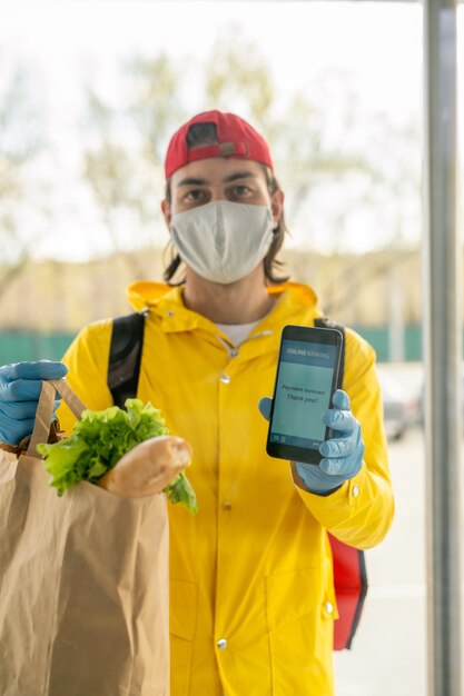 Foto retrato del repartidor con máscara y abrigo amarillo que muestra el mensaje en el teléfono inteligente mientras realiza la entrega sin contacto de productos