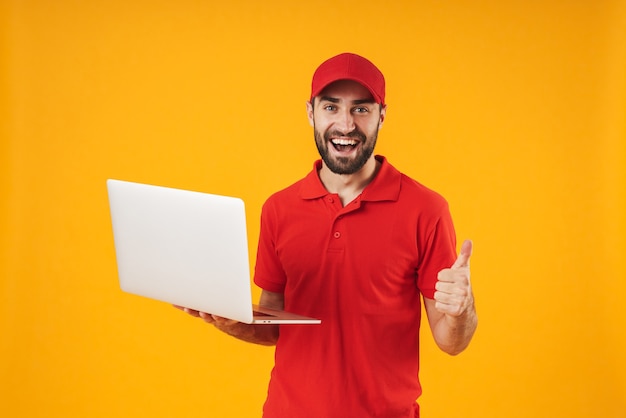 Retrato de repartidor positivo en camiseta roja y gorra sonriendo y sosteniendo un ordenador portátil plateado aislado sobre amarillo