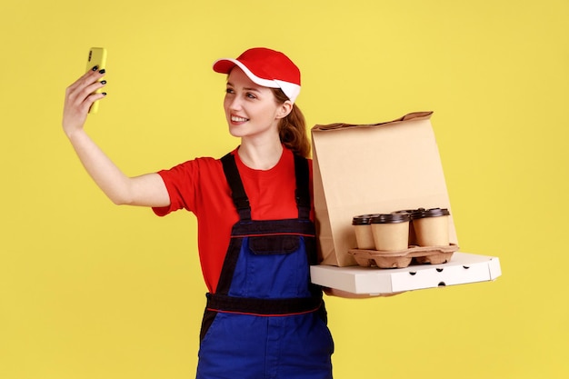 Retrato de una repartidora positiva sonriente sosteniendo cajas de café y pizza, tomando selfie o transmitiendo en vivo, usando overoles y gorra roja. Disparo de estudio interior aislado sobre fondo amarillo.