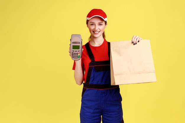 Retrato de una repartidora que sostiene un paquete de papel y una terminal, pago sin contacto, mirando la cámara con expresión feliz, usando overoles y gorra. Disparo de estudio interior aislado sobre fondo amarillo.