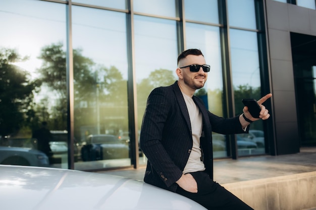 Foto retrato de un rico hombre de negocios con traje de pie cerca de su lujoso coche negro y usando un teléfono inteligente mientras lo sostiene en la mano