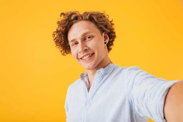 Retrato de rizado chico caucásico riendo y tomando selfie foto, aislado sobre fondo amarillo