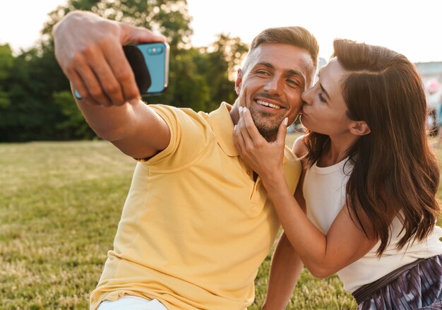 Retrato de romántica pareja adulta hombre y mujer besándose y tomando foto selfie en teléfono celular mientras está sentado en el césped en el parque