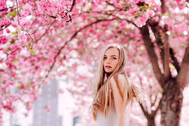 Retrato romântico da loira encantadora na parede de um jardim florescendo. árvore com flores cor de rosa. mulher loira de beleza com cabelos longos ao ar livre. flores de cerejeira. feche o retrato. sakura