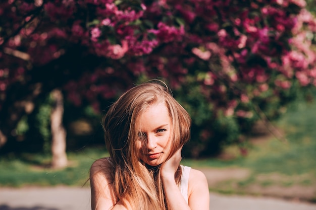 Retrato romântico da loira encantadora na parede de um jardim florescendo. Árvore com flores cor de rosa. Mulher loira de beleza com cabelos longos ao ar livre. Flores de cerejeira. Feche o retrato. Sakura