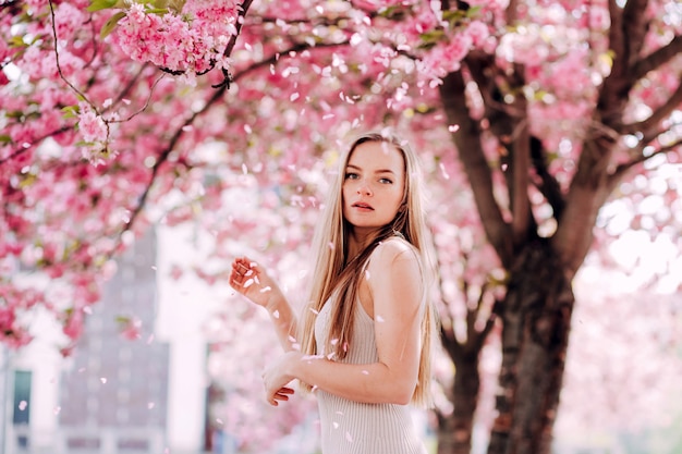 Retrato romântico da loira encantadora na parede de um jardim florescendo. Árvore com flores cor de rosa. Mulher loira de beleza com cabelos longos ao ar livre. Flores de cerejeira. Feche o retrato. Sakura