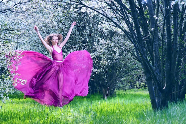 Retrato romântico da mulher de vestido carmesim arejado dançando entre as árvores florescendo