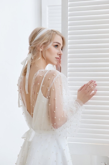 Retrato romántico de una mujer cerca de la ventana con un hermoso vestido largo blanco en casa. La chica es rubia con ojos azules y hermoso maquillaje en su rostro. Cosmética natural