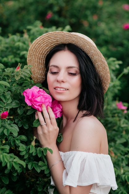 Retrato romántico de primer plano o encantadora mujer morena con sombrero de paja huele a flores en rosales