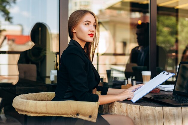 Retrato de rubia empresaria en café sosteniendo un documento y mirando soñadoramente al aire libre