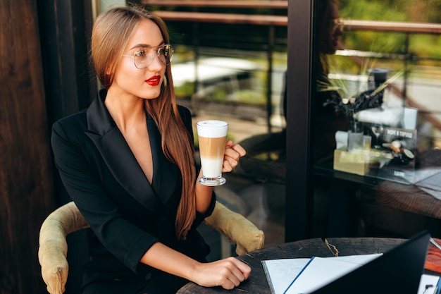 Retrato de rubia empresaria en copas sosteniendo una taza de café y mirando directamente