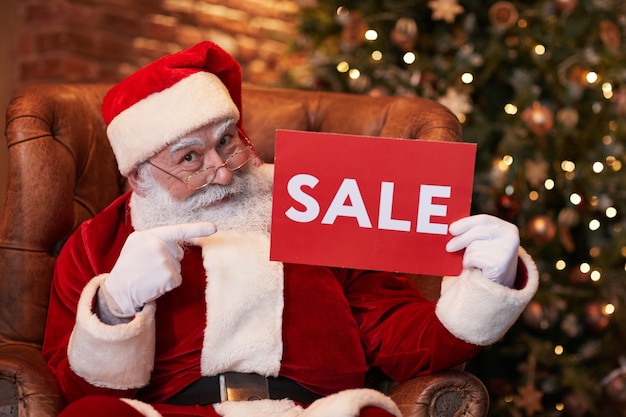 Retrato de santa claus barbudo positivo en anteojos sentado en un sillón y apuntando al banner de venta