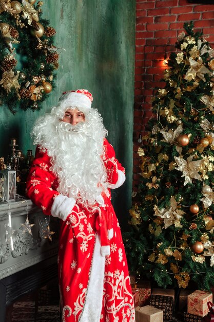 Retrato de un Santa Claus emocional con bolsa de regalos junto a la chimenea de Navidad en salón decorado con árbol de Navidad. Santa Claus viene y trae regalos por la noche. Copie el espacio para el sitio