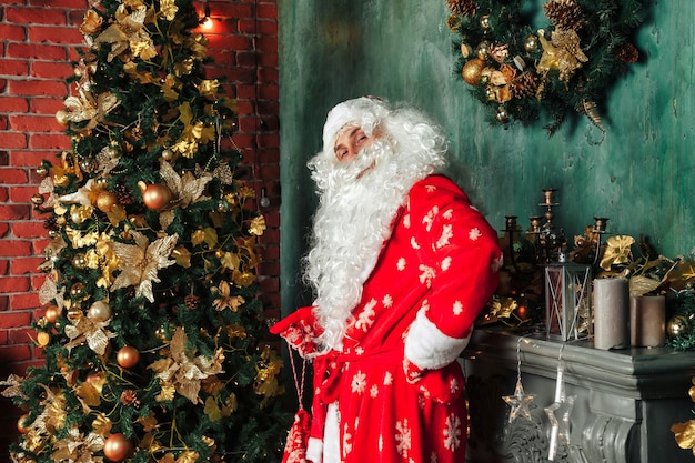 Retrato de un Santa Claus emocional con bolsa de regalos junto a la chimenea de Navidad en salón decorado con árbol de Navidad. Santa Claus viene y trae regalos por la noche. Copie el espacio para el sitio