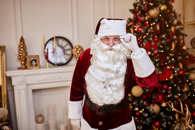 Retrato de Santa Claus con gafas cerca de la chimenea y árbol de Navidad con regalos.