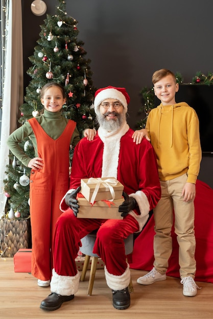 Retrato de santa claus sonriente en traje rojo sentado con regalos contra el árbol de navidad mientras posa ...