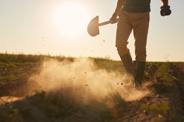 Retrato de sección baja de un trabajador masculino irreconocible que sopla nubes de polvo de botas de goma y sostiene una pala mientras camina por el campo de la plantación en la luz del atardecer, espacio de copia