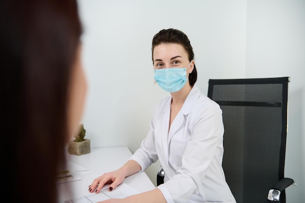 Retrato seguro de una doctora morena caucásica con máscara médica mirando la cámara mientras consulta a un paciente en el moderno consultorio médico minimalista en una clínica con interior blanco
