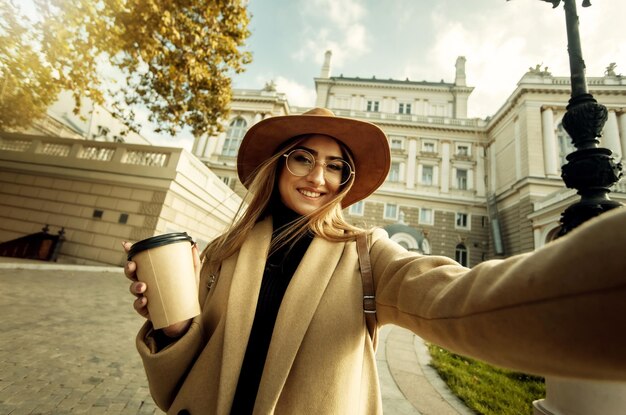 Retrato selfie de una joven turista con estilo vestida con un abrigo y un sombrero sobre un fondo de arquitectura urbana europea. Concepto de turismo y vacaciones