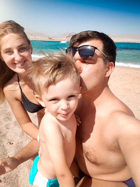 Retrato de Selfie de padres besando a su hijo pequeño en la playa del mar en un día soleado y ventoso. Familia relajándose y pasando un buen rato durante las vacaciones de verano.