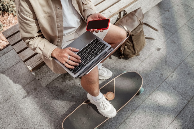 Retrato sem rosto de jovem patinador sentado na rua, trabalhando no laptop, segurando o telefone móvel com longboard e mochila. conceito de trabalho remoto. cara de hipster milenar. mãos digitando de cima