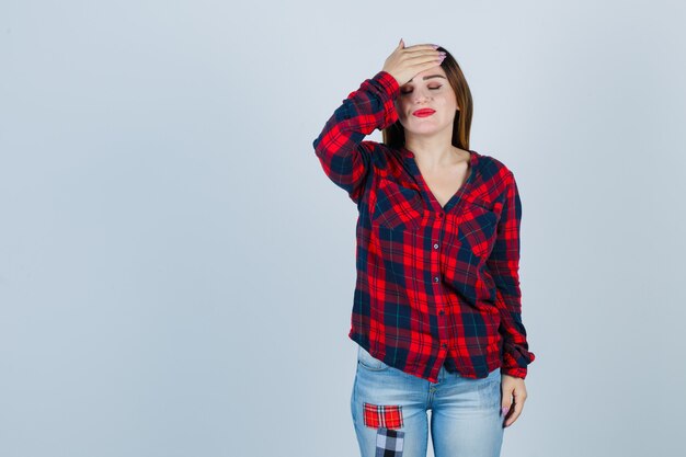 Retrato de señorita con la mano en la frente mientras cierra los ojos en camisa a cuadros, jeans y mira arrepentida vista frontal