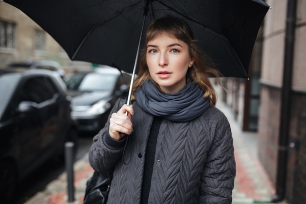 Retrato de señorita de pie en la calle con paraguas negro en la mano y pensativamente