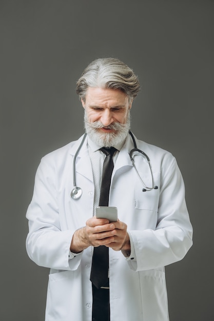 Retrato del sensor médico en la pared gris. El señor doctor canoso vestido con bata blanca hablando por teléfono.