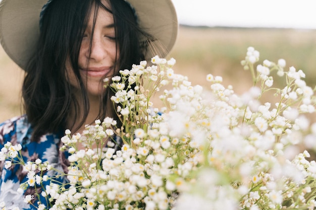 Retrato sensual da menina bonita com grande buquê de margaridas no campo ventoso verão no campo Bela jovem de vestido vintage azul e chapéu segurando flores silvestres brancas no Prado