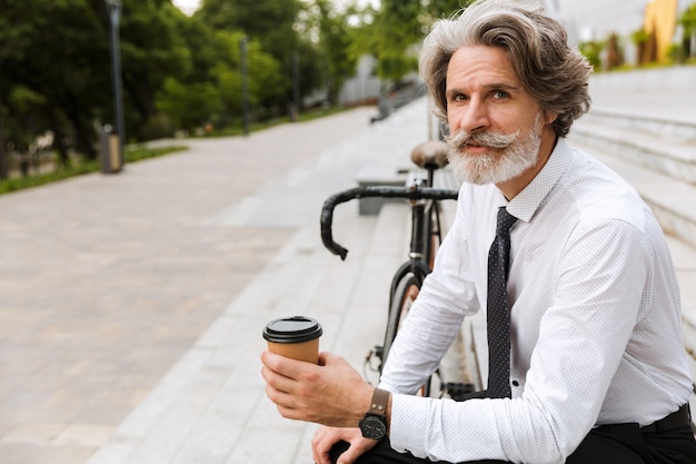Retrato de serio empresario barbudo en ropa formal bebiendo café para llevar mientras está sentado en un banco con bicicleta al aire libre