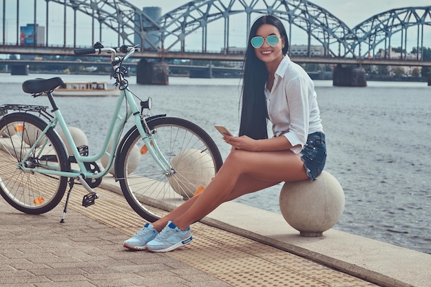 Retrato de una sexy morena sonriente vestida con blusa y pantalones cortos con gafas de sol, sentada en un muelle contra el puente, sosteniendo un teléfono inteligente, relájese después de andar en bicicleta.