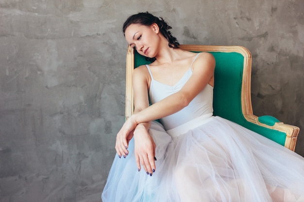 Retrato sincero de bailarina de ballet bailarina en un hermoso vestido azul claro, falda tutu posando sentada en una silla vinage en estudio loft