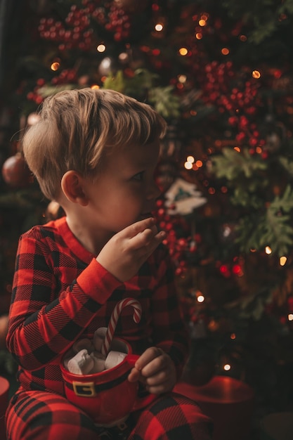 Retrato sincero niño feliz en pijama a cuadros rojos sostenga la taza de Navidad con malvaviscos y bastón de caramelo