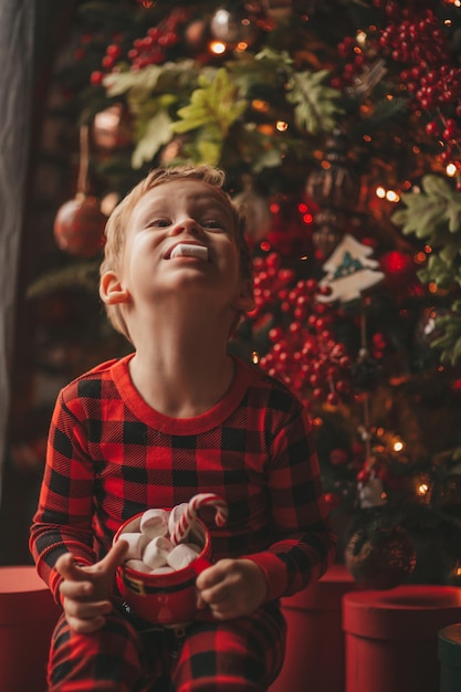 Retrato sincero niño feliz en pijama a cuadros rojos sostenga la taza de Navidad con malvaviscos y bastón de caramelo