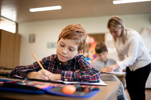 Retrato de sonriente alumno caucásico prueba de escritura en su aula y profesor en segundo plano.