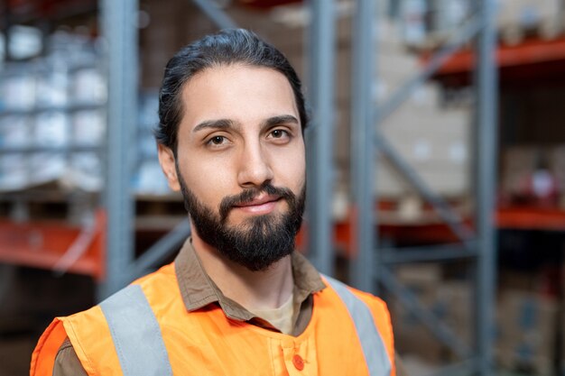 Retrato de sonriente apuesto joven trabajador de raza mixta en chaleco naranja de pie contra estantes metálicos en el almacén