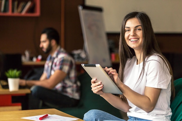 Retrato de una sonriente y atractiva empresaria morena que trabaja con una tableta en una oficina moderna sentada cerca de la mesa