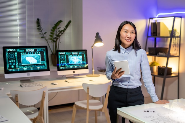 Retrato de sonriente atractivo diseñador de automóviles asiáticos en camisa despojada sosteniendo tableta digital en la oficina moderna con computadoras