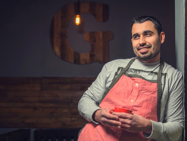 Retrato de un sonriente baristas barbudos en un delantal rojo con una taza en las manos en un acogedor café.