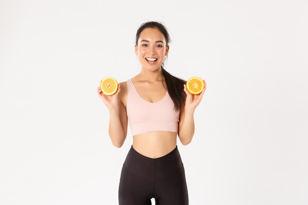 Retrato de sonriente chica asiática sana y delgada consejos comer alimentos saludables para el desayuno, ganar energía para hacer ejercicio, sostener dos mitades de naranja.